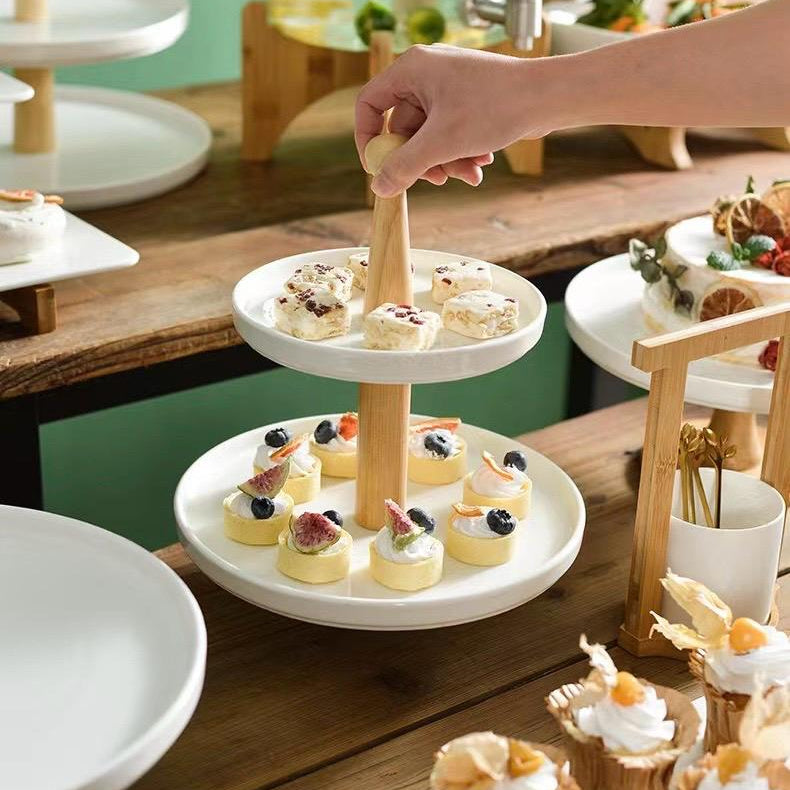 A person's hand is seen placing a dessert on a white porcelain serving tray with a bamboo stand, which is part of a larger serving setup with various other trays and food items in the background.