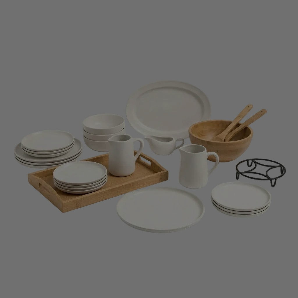 White plates, bowls and oval platter with white pitchers, a bamboo serving tray, a wooden salad bowl with servers and a black metal trivet