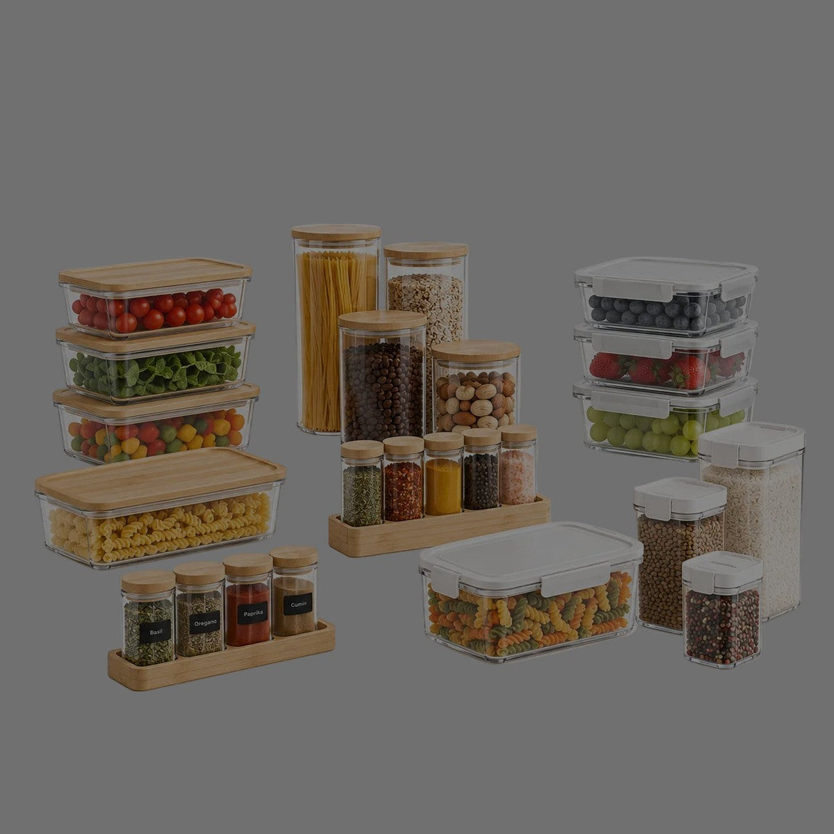 Glass containers with bamboo lids filled with vegetables and pasta, tall glass jars, spice jars on bamboo trays and white-lid airtight containers with grains and fruit