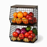 Two stacked black wire baskets with wooden tops holding oranges, apples, onions, tomatoes, peppers and potatoes