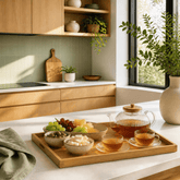 A styled kitchen scene with a wooden serving tray holding a glass teapot, two cups of tea, and small bowls of fruit and cheese on a white countertop. The background shows light wood cabinets, a pale green tiled backsplash, and a window with greenery.