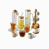 A tea and coffee serving set on a white background, including two glass carafes with bamboo lids, two clear glass cups with amber liquid, two white floral mugs, a glass cup with a coaster, a mug with a coaster, a cup on a stand, and a wooden mug rack.