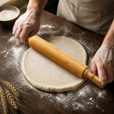 Hands rolling out pizza dough on a floured dark wooden table with a bamboo rolling pin
