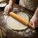Hands rolling out pizza dough on a floured dark wooden table with a bamboo rolling pin