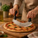Hands cutting a margherita pizza with basil on a wooden board using a curved double-handle pizza cutter