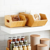 Two bamboo bins with cut-out side handles on a white shelf holding walnuts and bagged cashews, above a rack of spice jars and tan canisters
