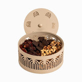 Round beige metal bowl with arch cut-outs and open lid, holding dates, dark chocolate and mixed nuts