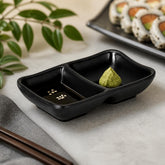 Black matte divided sauce dish with soy sauce and sesame in one section and wasabi in the other next to sushi rolls