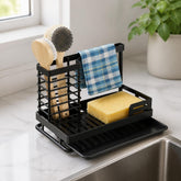 Black sink caddy holding 2 wooden dish brushes, a yellow sponge and a checkered towel next to a kitchen sink