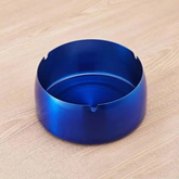 Blue stainless steel round ashtray with cigarette notches on a light wooden table