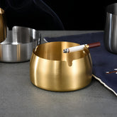Gold ashtray with a lit cigarette on a grey table next to silver ashtrays and a navy cloth