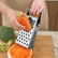 Hand grating a carrot on a stainless steel box grater over a wooden cutting board, with shredded carrot on a plate and broccoli beside it