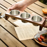 Hands lifting a wooden stand with 3 empty steel sauce cups over an outdoor table