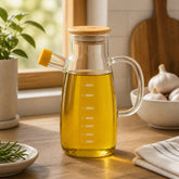 Glass olive oil jug full of olive oil on a wooden kitchen counter by a sunny window with garlic and rosemary