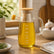 Glass olive oil jug full of olive oil on a wooden kitchen counter by a sunny window with garlic and rosemary
