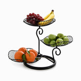Black 3-tier metal fruit stand with leaf-shaped wire baskets holding grapes, bananas, green apples and oranges