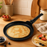 Golden crepe in a granite crepe pan on a wooden counter next to folded crepes with strawberries and blueberries