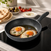 Granite frying pan with two fried eggs, sumac and parsley on an induction cooktop next to pita, olives and labneh