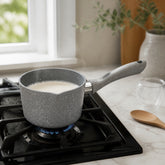 Grey granite saucepan warming milk on a gas stove next to a marble counter with a wooden spoon