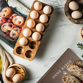 Rectangular wooden egg tray with eggs next to muffins, bread and a bamboo steamer