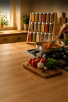 A stainless steel frying pan sits on a gas stovetop in a warm, rustic kitchen setting, with a hand holding a wooden spoon and stirring the food. In the foreground, fresh tomatoes, garlic, and herbs rest on a wooden cutting board, with a spice rack filled with jars in the background.