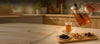 A hand pours amber tea from a glass teapot with an infuser into a small clear glass cup on a wooden tray, set on a table in a warm, softly blurred kitchen setting.