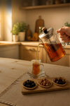 A hand holds a clear glass teapot with amber tea and an infuser over a small clear glass cup on a table. A wooden tray with small dishes of olives sits nearby in a warm, softly lit kitchen setting.
