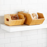 Small and large bamboo bins with side handles on a white shelf, one holding walnuts in a clear container and the other holding bagged cashews