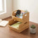 Two stacked bamboo storage bins on a wooden desk, the top one holding scissors, a remote and a jar of paper clips and the bottom one holding a green notebook, beside a small succulent