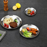 Three stainless steel plates in different sizes on a dark slate table holding spaghetti with steak, olives with tomatoes, and chicken wings with lettuce, beside a pepper mill and lemons