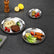 Three stainless steel plates in different sizes on a dark slate table holding spaghetti with steak, olives with tomatoes, and chicken wings with lettuce, beside a pepper mill and lemons