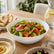 White glass bowl with wavy rim filled with fattoush salad on a dining table with pita, hummus and olives
